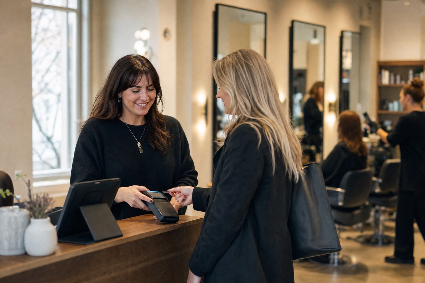 Friseurin am Empfangstresen eines Salons rechnet mit einer Kundin ab, im Hintergrund Behandlungsplätze (Symbolbild)